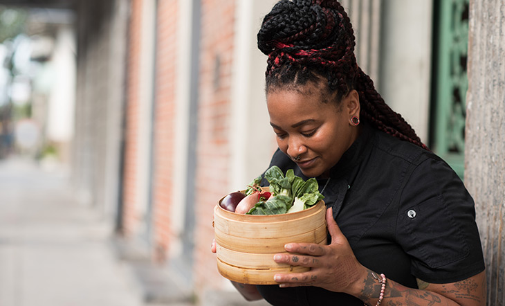 Myisha Mastersson in black chef's jacket holding and looking at steamer basket of vegetables photo by Kayla Madden Photography 