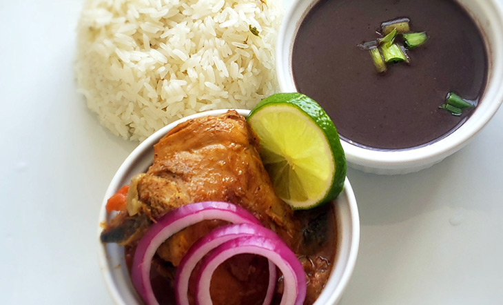 Chicken stew in a white container with a cup of soupy beans and a mound of rice on the side photo by Fritz Edmonis