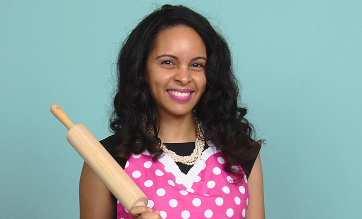 Photo of founder of The Pink Bakery Nubian Simmons. She is wearing a polka dotted apron and pearls and is holding a rolling pin. 