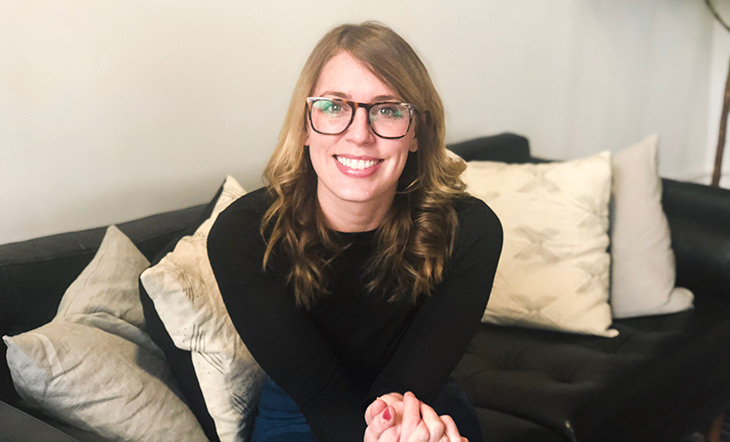 Headshot of Elizabeth Tilton, a blond woman in glasses wearing a black shirt and jeans sitting on a black sofa with cream colored and grey pillows 