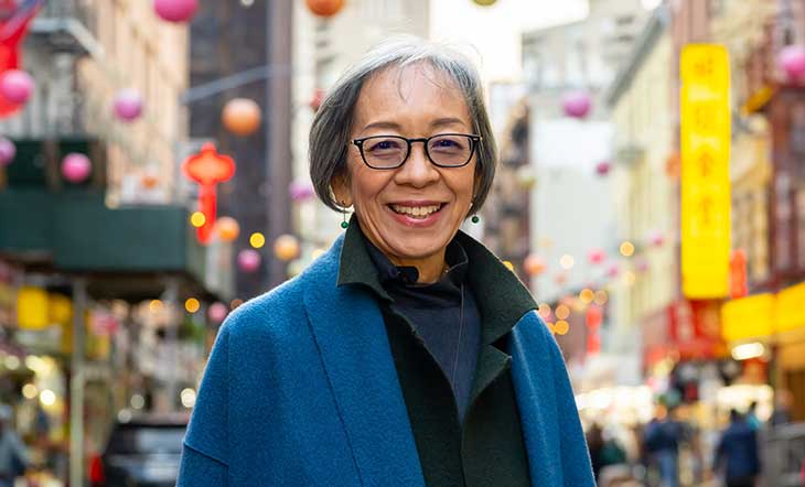 Photo of Grace Young in blue coat and blue and green shirt layers with paper lanterns and Chinatown business signs behind her photo by Zabrina Deng